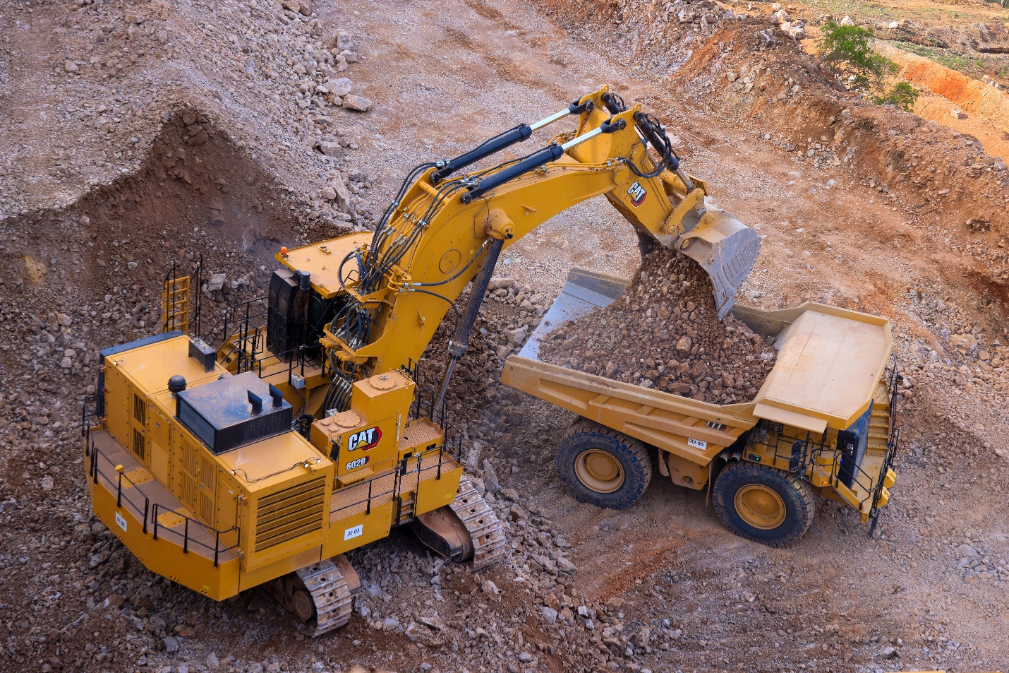 The image shows a large, yellow excavator (CAT 6020 EXC-01 CAM-05) in action, digging and moving earthy, unpaved soil.
AI-generated content may be incorrect.
