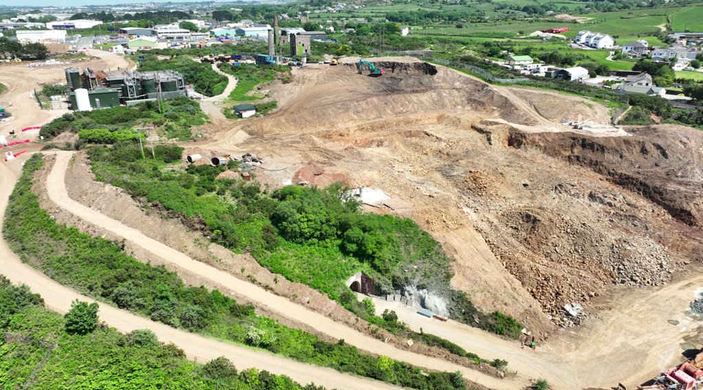 The image depicts a large, barren construction site surrounded by greenery, with visible machinery and a few buildings in the distance.

AI-generated content may be incorrect.