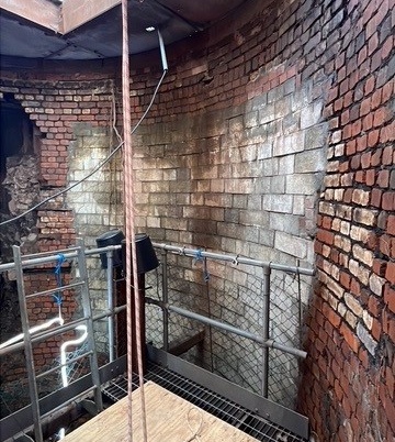 The image shows a brick wall being demolished, with workers wearing safety gear and equipment set up around the structure.

AI-generated content may be incorrect.
