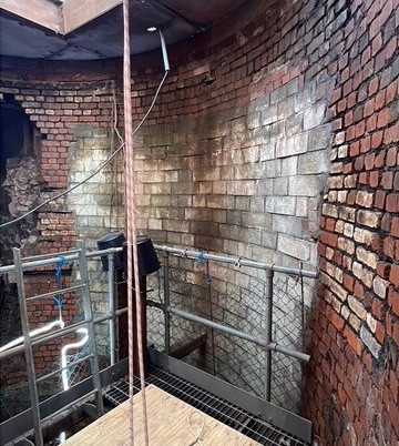The image shows a brick wall being demolished, with workers wearing safety gear and equipment set up around the structure.
AI-generated content may be incorrect.